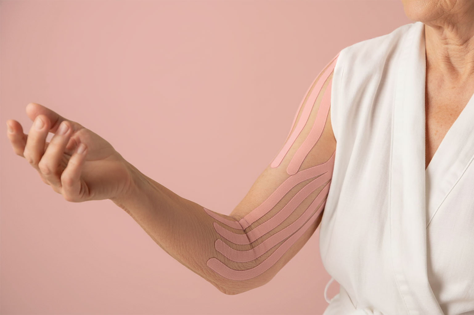 Ältere Person mit rosafarbenem, sanftem Kinesiotape entlang des Arms und Ellenbogens, vor rosa Hintergrund, Fokus auf die hautschonende Anlage.
